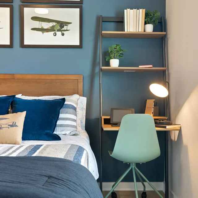 A cozy bedroom featuring a wooden bed with blue and white striped bedding, adorned with decorative pillows. Above the bed, there are three framed airplane prints. To the side, there’s a desk with a green chair, a lamp, and some decorative plants on shelves against a blue wall. Natural light pours in through a window with white blinds.