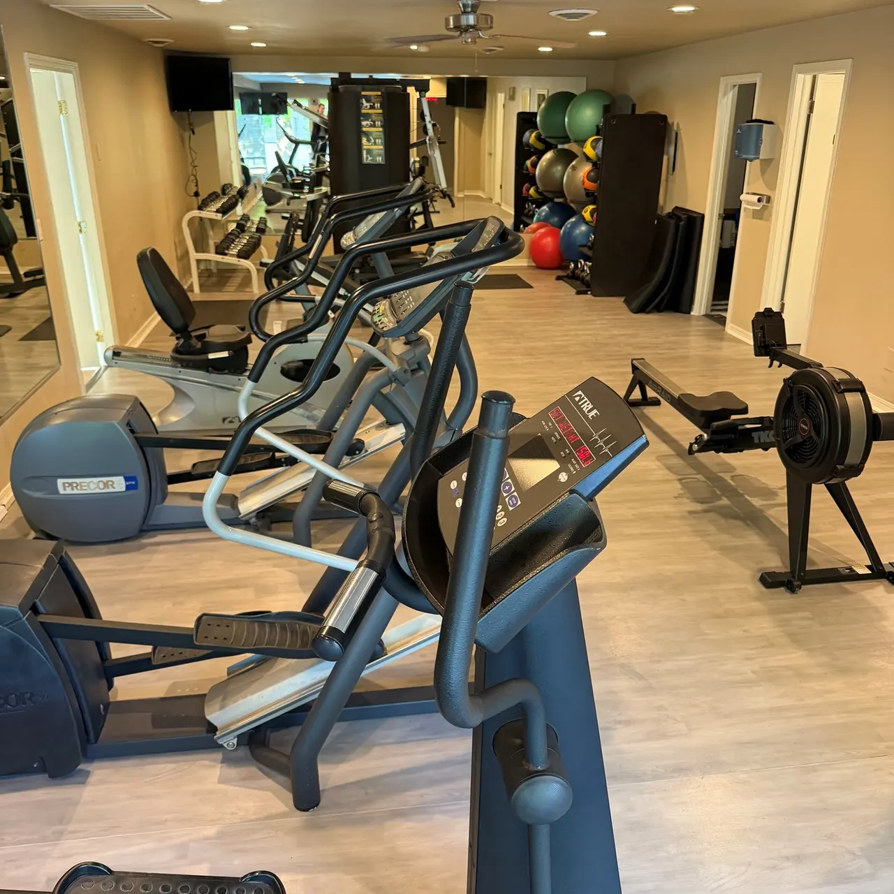 A modern gym interior featuring various exercise machines including elliptical trainers, a rowing machine, and an assortment of exercise balls in the background.