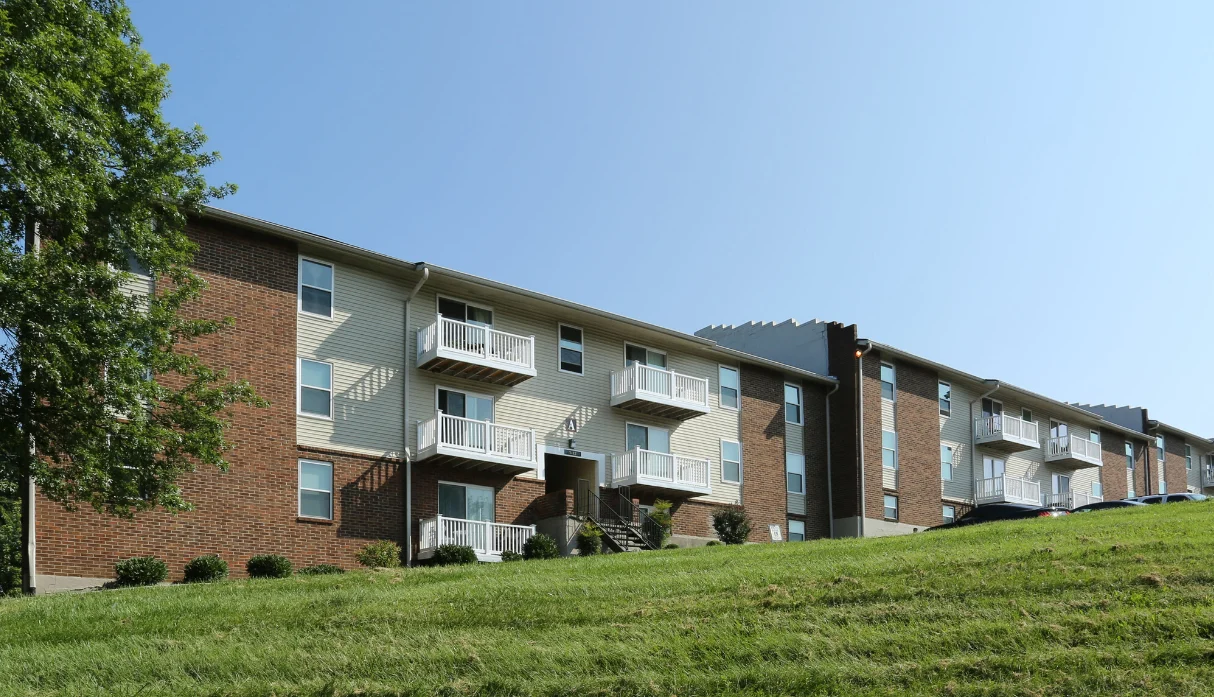 A two-story apartment building with brick and siding exterior, featuring balconies and a grassy area in front, under a clear blue sky.