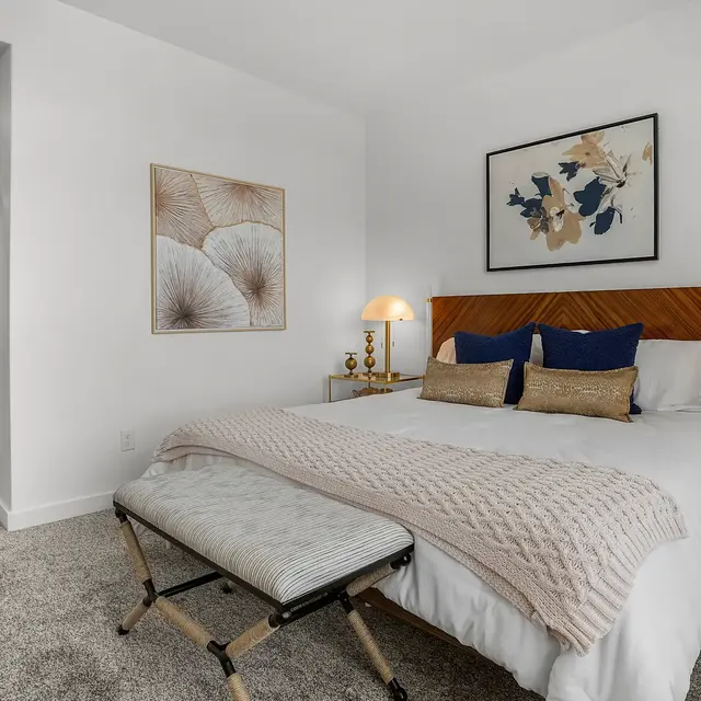 A modern bedroom featuring a neatly made bed with blue and beige pillows, decorative throw, and stylish side tables with lamps. The room is well-lit with a light carpet and artwork on the wall.