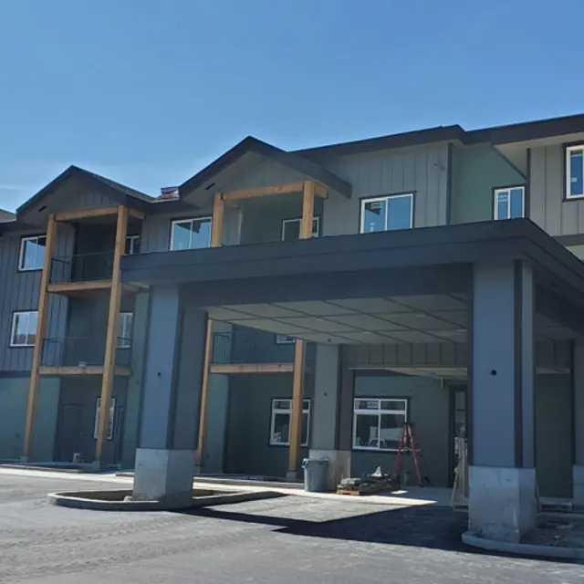 A newly constructed apartment complex featuring a modern design with a covered entrance. The building is two stories high, has a combination of green and wooden paneling, and large windows. The foreground includes a parking area.