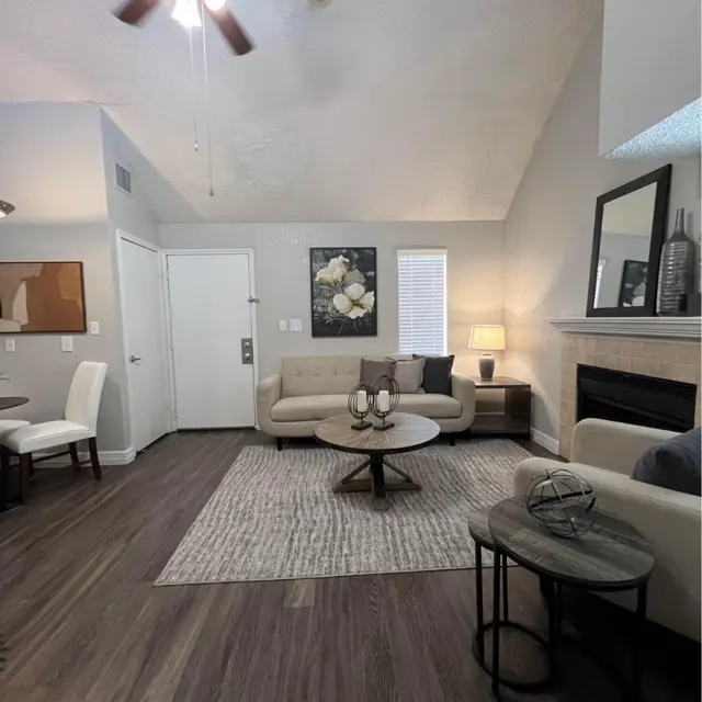 A modern living room featuring a beige sofa, a round coffee table, and a decorative rug on a dark wooden floor. A wall-mounted mirror and a themed artwork are visible, alongside a small side table and a ceiling fan.