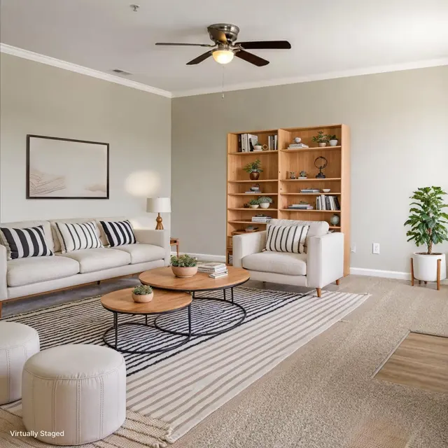 A modern living room featuring a light-colored sofa with striped cushions, a round wooden coffee table, and a bookshelf with decorative items. There are two ottomans and a potted plant in the corner, all set against a neutral wall with a ceiling fan above.