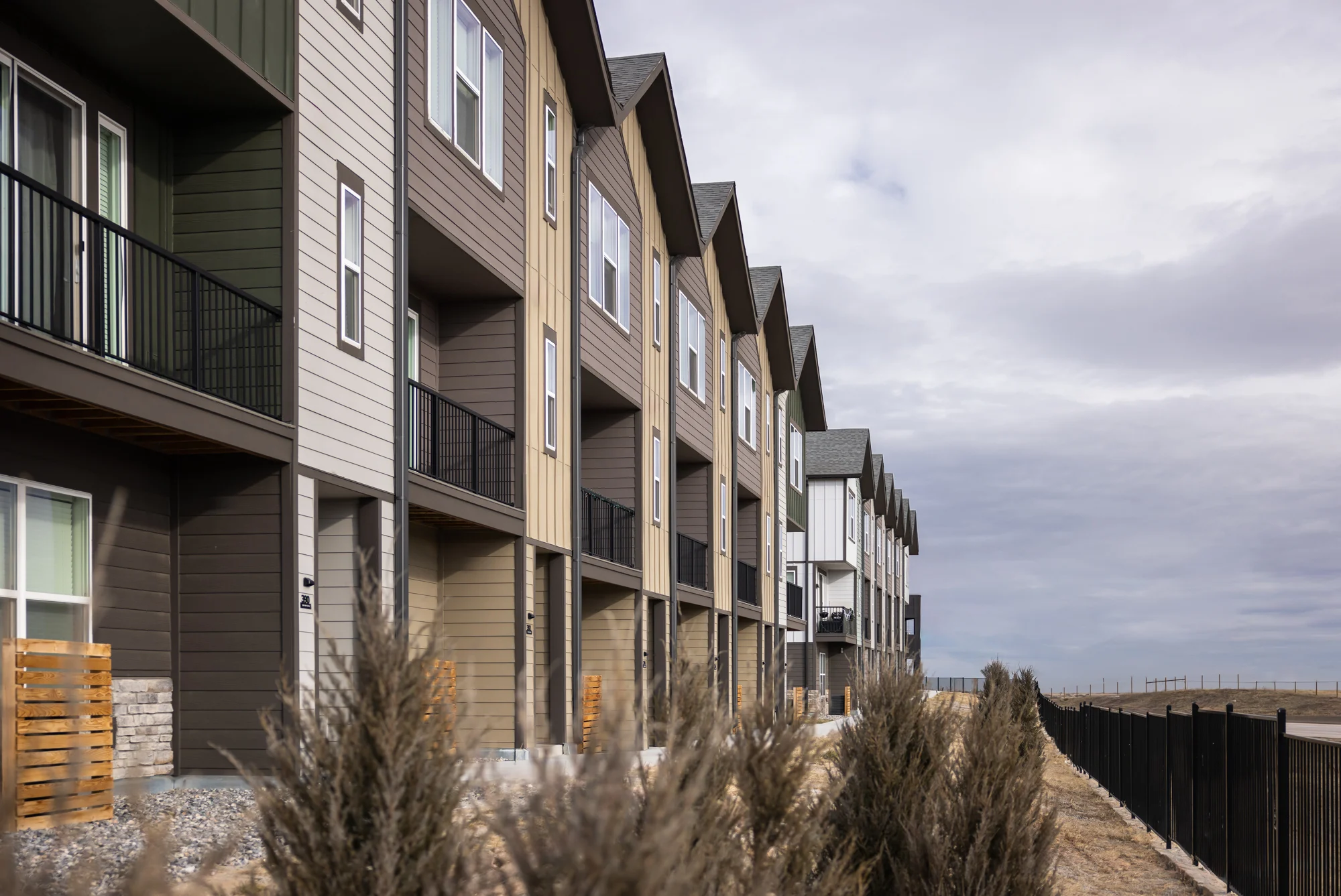 A row of modern townhouses with varying colors and features, set against a cloudy sky. The buildings have balconies and are bordered by low shrubs and fencing.