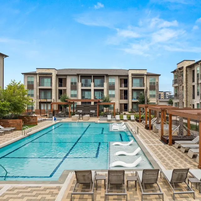 A modern apartment complex featuring a spacious outdoor swimming pool, surrounded by lounge chairs and shaded areas. The buildings are contemporary in design with greenery around.