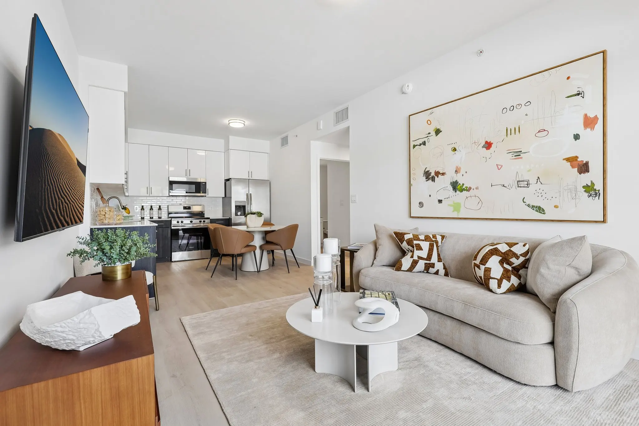 A modern living room featuring a comfortable sofa with decorative cushions, a round coffee table, a flat-screen TV on the wall, and a visible kitchen area in the background.