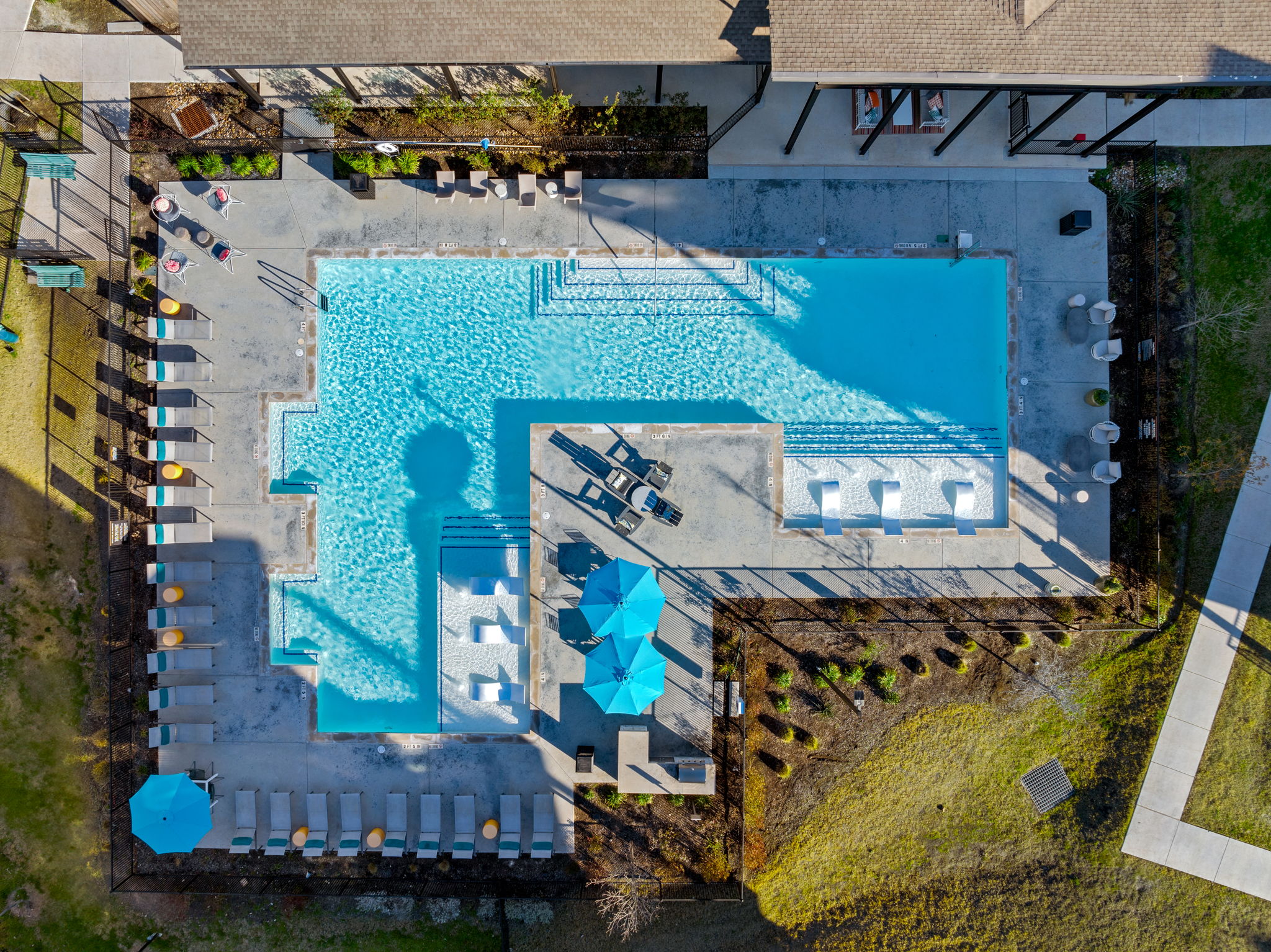 Aerial view of a rectangular swimming pool surrounded by lounge chairs and umbrellas, with landscaped areas and pathways nearby.