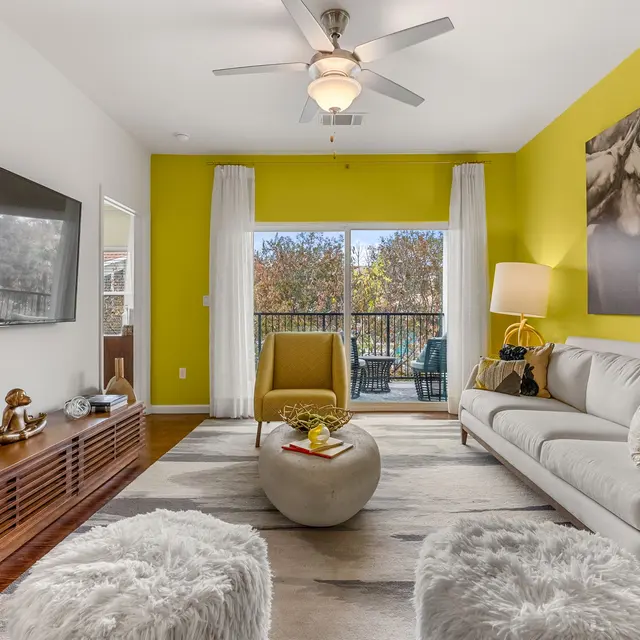 A contemporary living room featuring a bright yellow accent wall, large windows with sheer curtains, and a comfortable white sofa. The room includes a flat-screen TV, a stylish coffee table, and decorative items on a wooden media console. In the background, a balcony with outdoor furniture is visible.