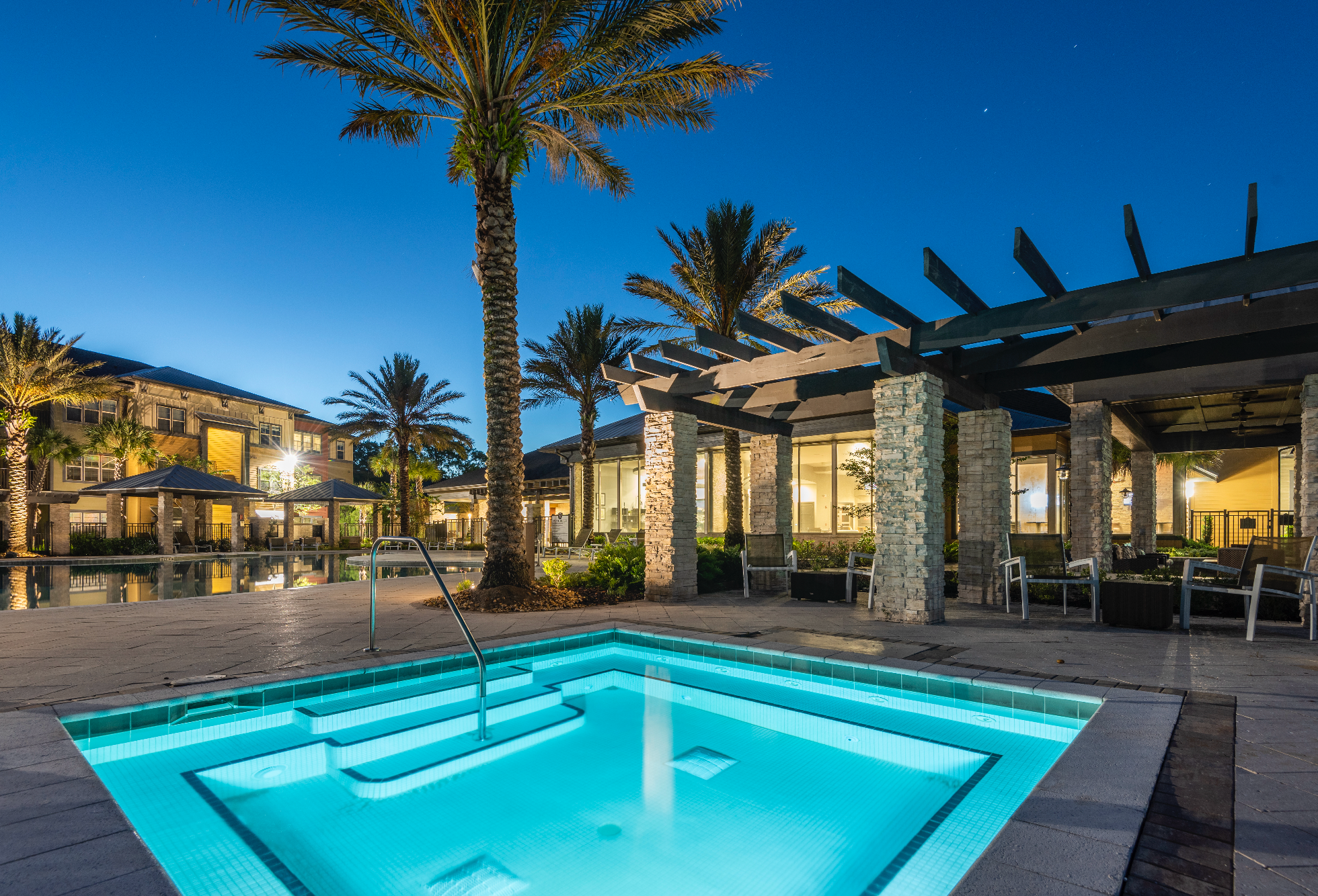 An inviting pool with a stair entry, surrounded by palm trees, at dusk. The pool is illuminated with a gentle blue light, and nearby covered seating areas are visible. In the background, elegant buildings glow warm from their lights.