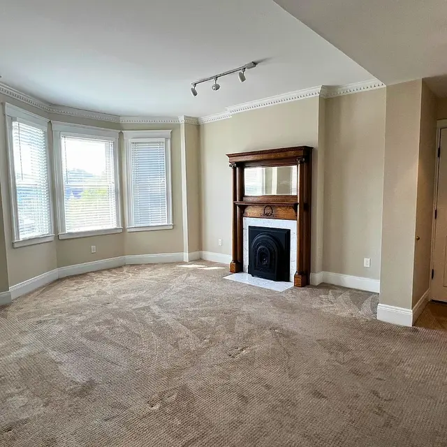 A spacious empty living room with beige carpet, featuring bay windows on one side and a decorative fireplace on the opposite wall, with natural light illuminating the space.