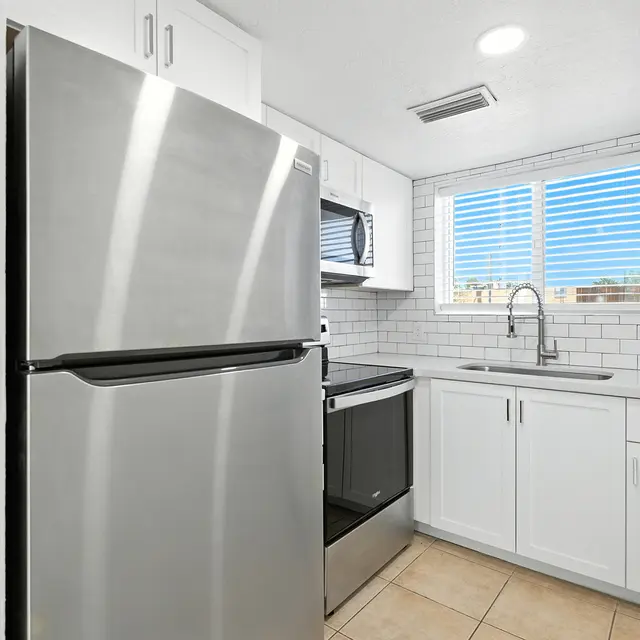A modern kitchen featuring stainless steel appliances including a refrigerator, microwave, and oven. Bright white cabinetry and tiled backsplash emphasize a clean aesthetic. A window above the sink allows natural light to illuminate the space.