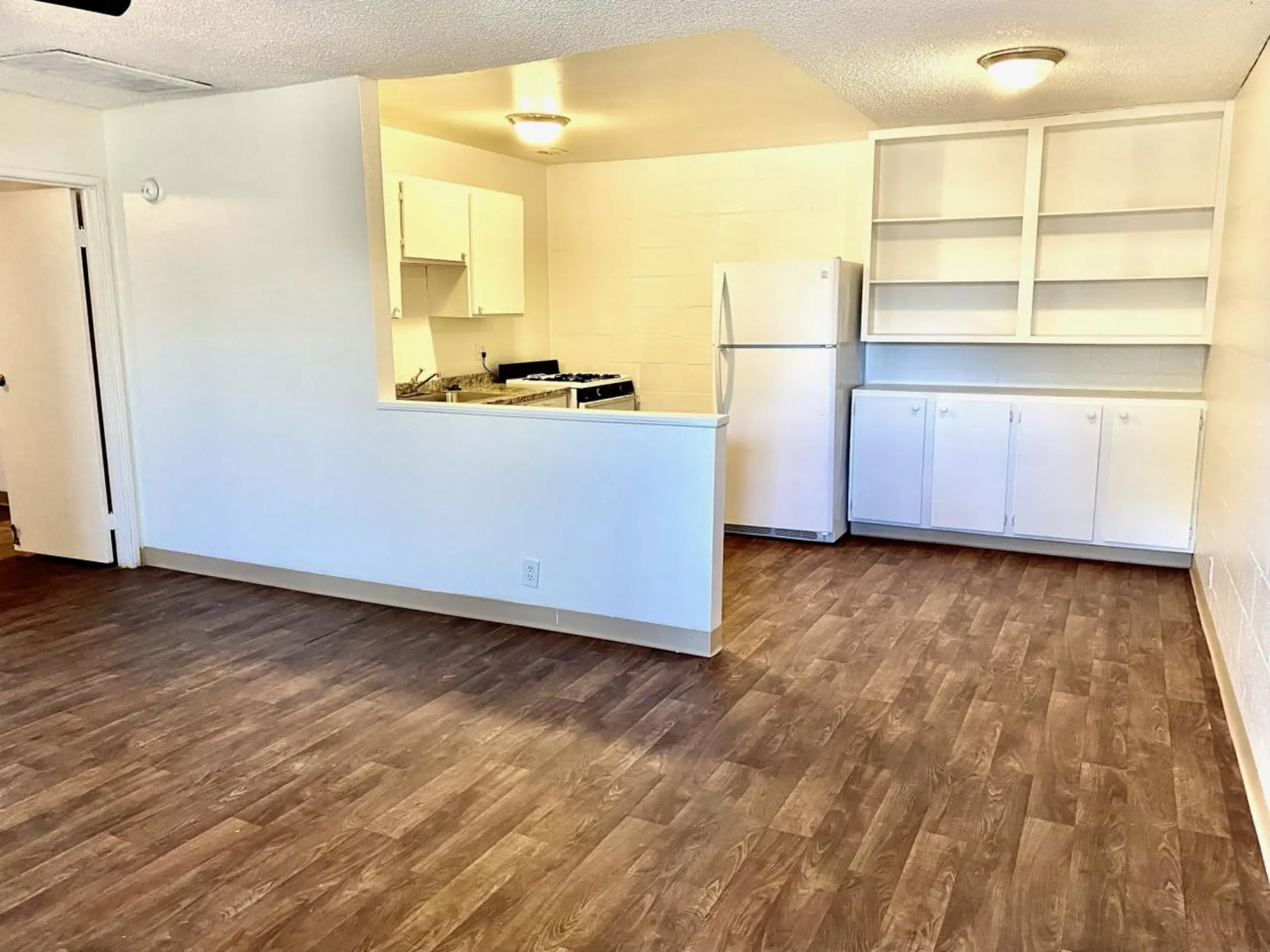 Open living area beside a kitchen with white cabinets, refrigerator and shelving.