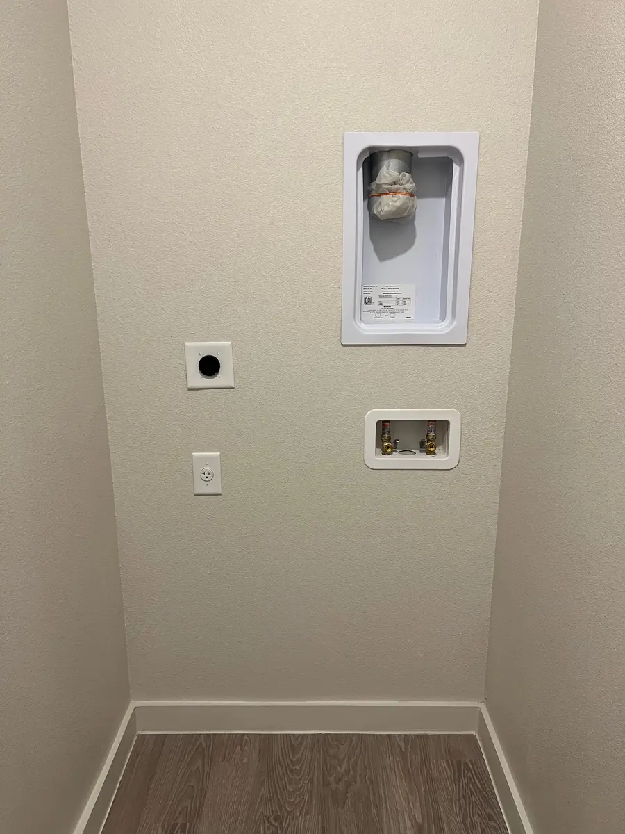 A wall section showing a washing machine hookup area in a minimalist design. There are connections for water and an electrical outlet, situated on a beige wall with a light wooden floor.