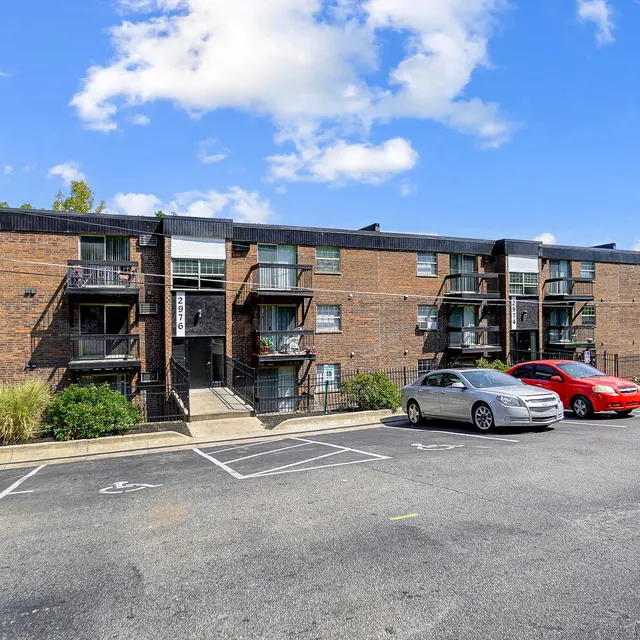 A view of a brick apartment complex with multiple stories and balconies. The foreground has several parked cars and a motorcycle. The sky is partly cloudy with a few trees in the background.