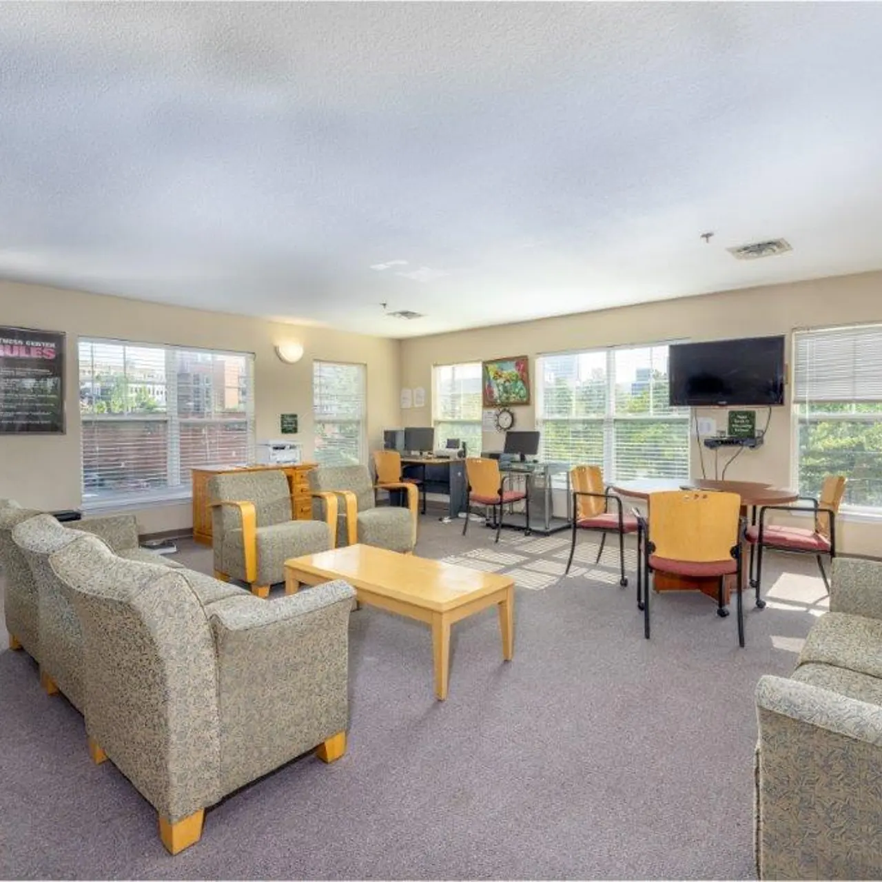 A bright and open community lounge area featuring upholstered seating, tables, and computers. Large windows provide natural light.