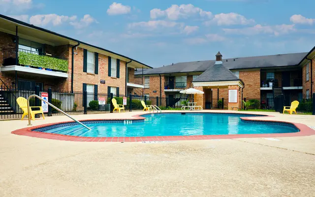 A swimming pool located in an apartment complex, featuring a shallow end with a slide, surrounded by sun loungers and brick buildings under a clear blue sky.