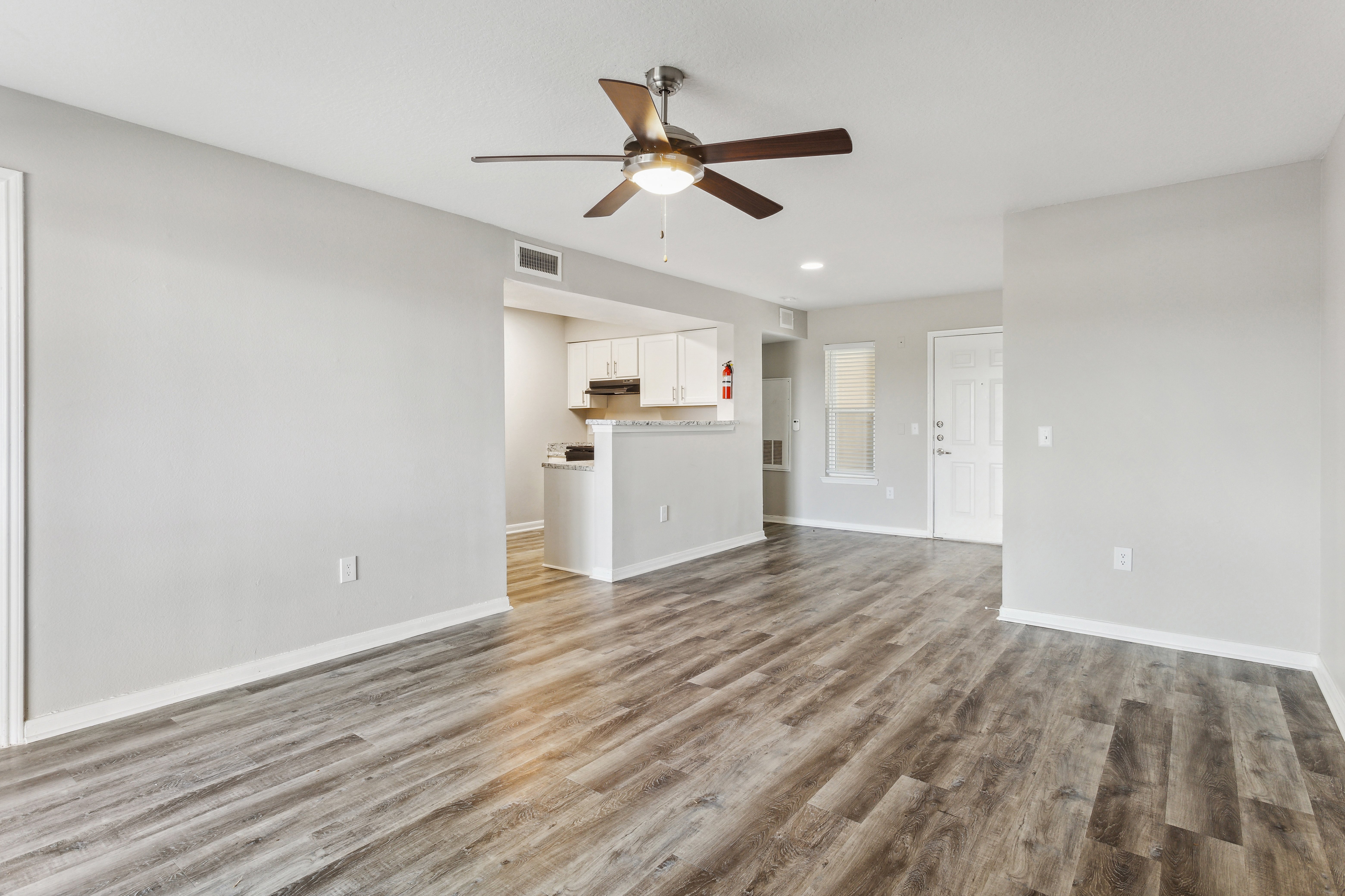 Spacious living area of a modern apartment featuring a ceiling fan, laminate flooring, and an open layout that connects to a kitchen area.