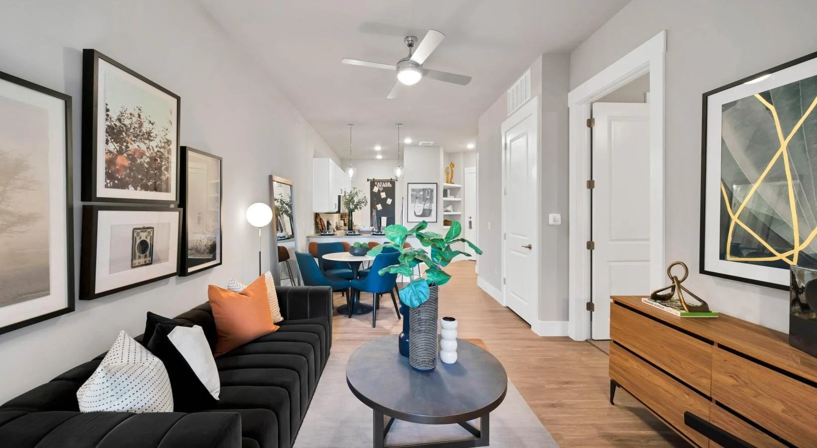 A stylish modern living room with a black sofa, coffee table, and decorative elements. The walls are adorned with framed artwork, and there are plants adding greenery. The space features a dining area and a bright, open feel with a fan installed.