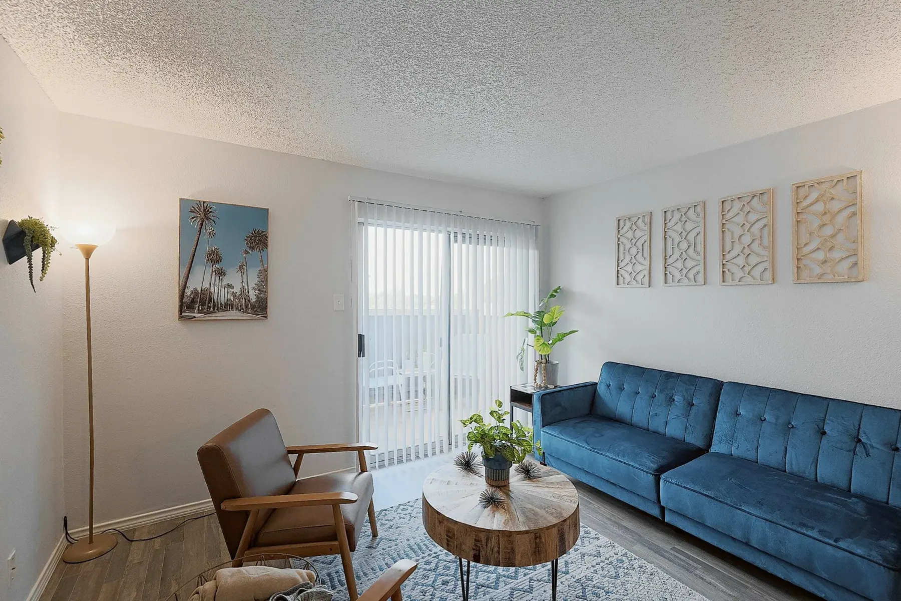 Cozy living room with a blue sofa, round coffee table, plants, and decorative wall art. Bright with natural light from sliding doors.
