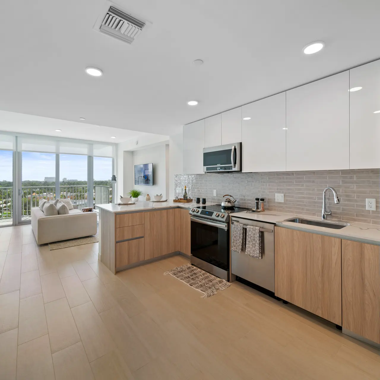A modern kitchen with light wood cabinetry and stainless steel appliances, featuring a spacious layout and large windows overlooking a balcony.
