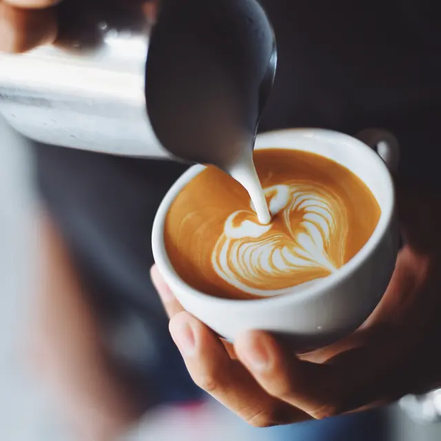 A person pouring milk into a cup of coffee, creating a heart-shaped latte art design.
