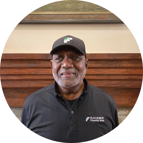 A smiling man wearing a black polo shirt and cap with a logo, standing in front of a wooden backdrop.