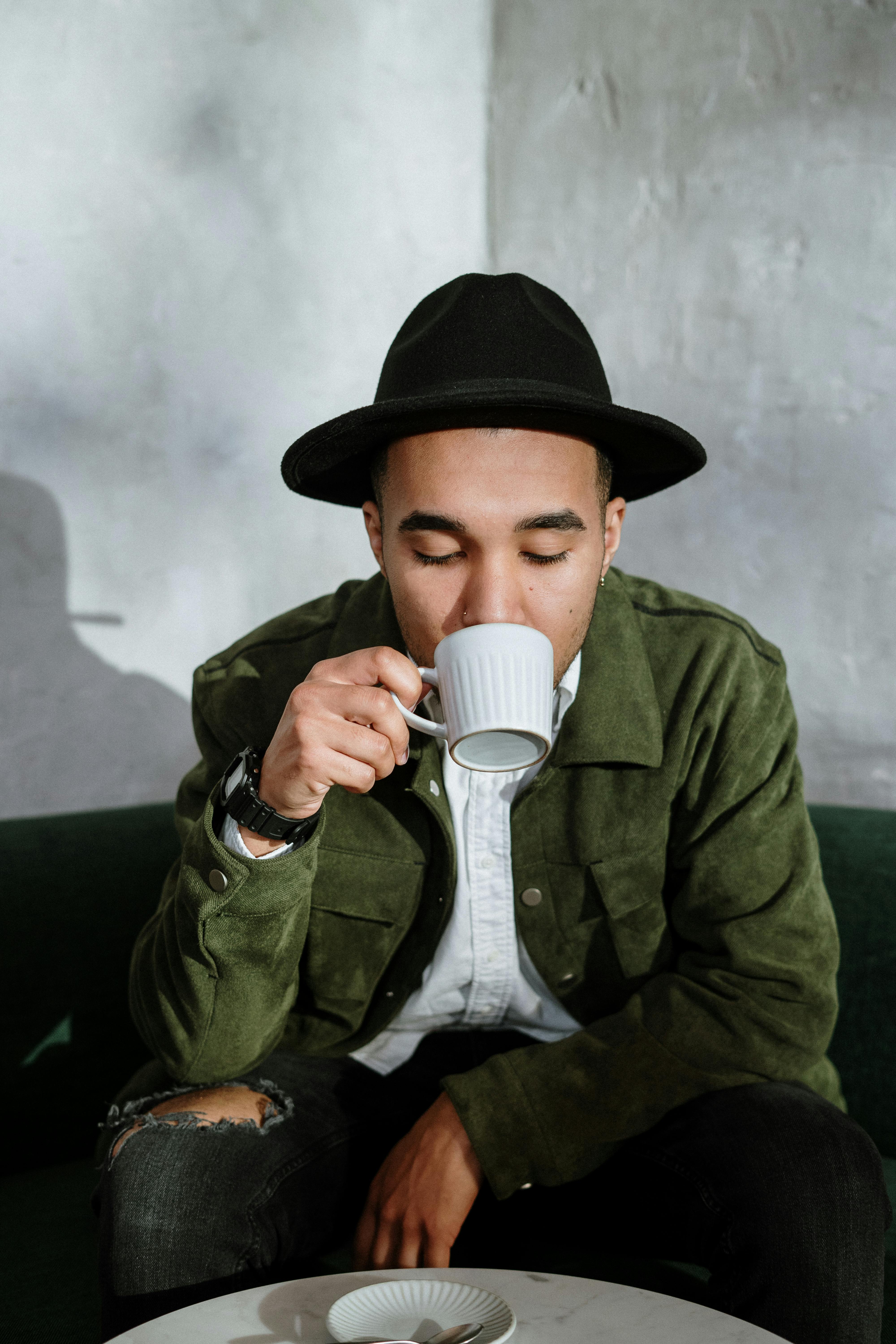 Young Man Sipping Coffee A young man wearing a green jacket and black hat is sipping from a white coffee cup while seated on a green couch.