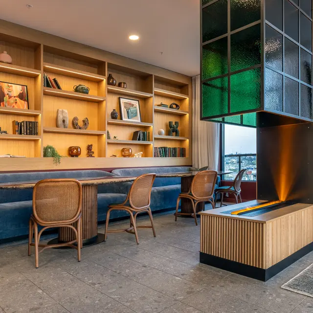 A modern cozy living space featuring a blue sofa, wooden shelves filled with decorative items, and a stylish room divider with a glass feature. There are four rattan chairs around a small table, with natural light coming through large windows.