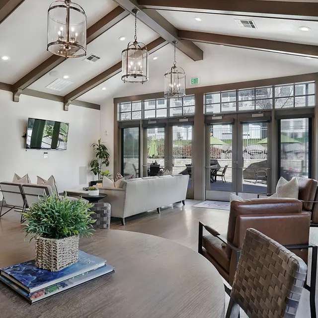 A spacious modern living room featuring high ceilings with wooden beams, large windows, and comfortable seating arrangements. A coffee table adorned with a plant and books is centrally placed, while stylish pendant lights hang above.
