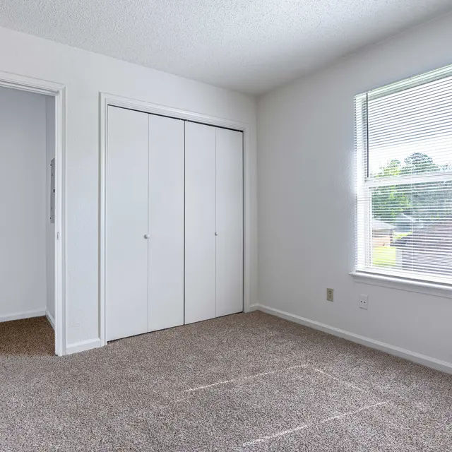 A clean, empty bedroom featuring beige carpet, white walls, and a window with blinds.