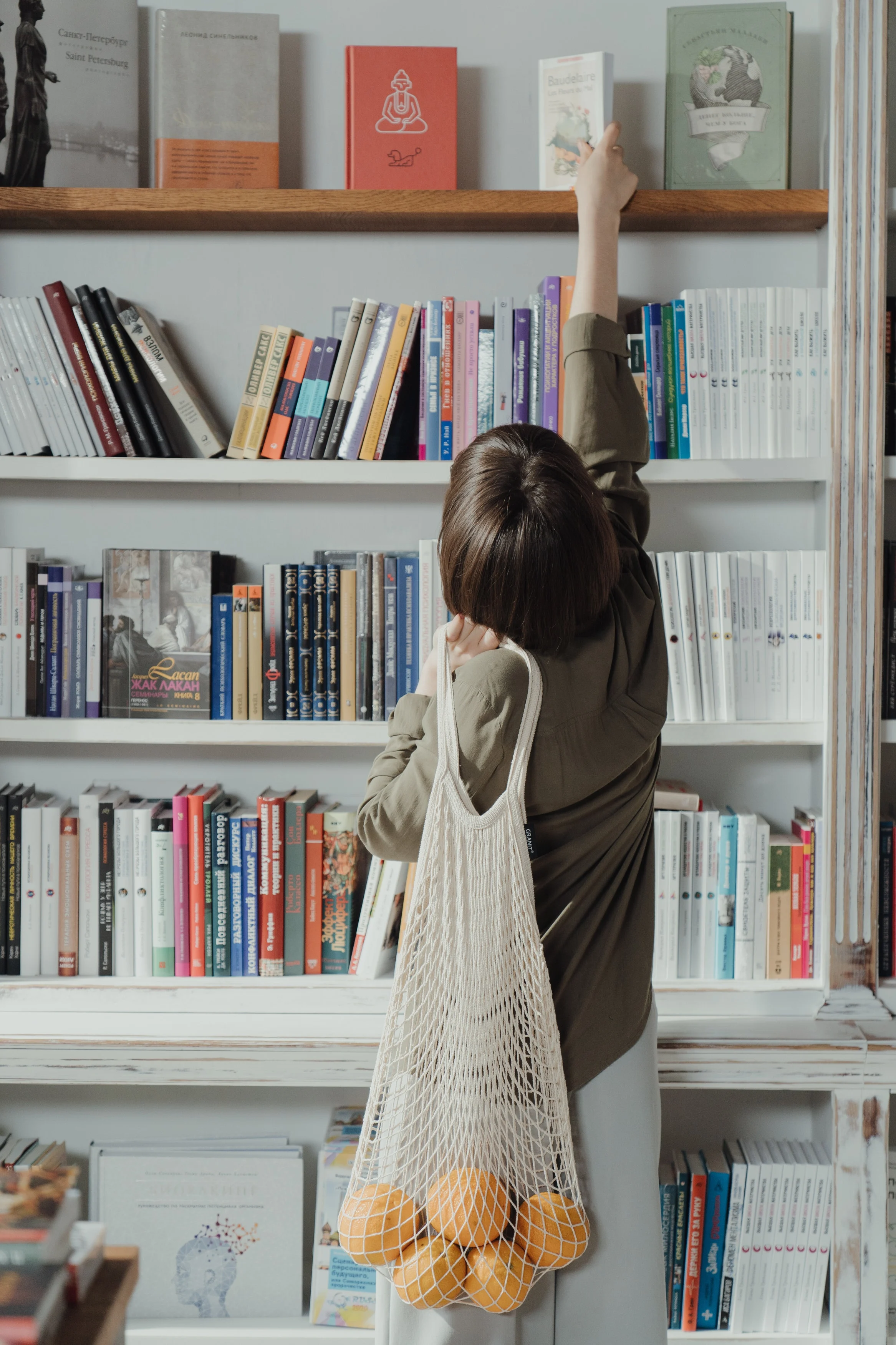 A person reaching for a book on a shelf in a library, with a net bag containing oranges slung over their shoulder.