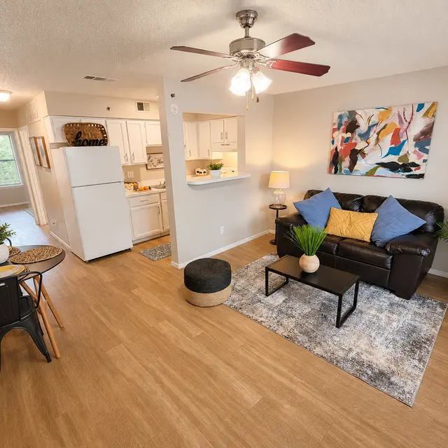 A modern living room with a brown couch, colorful artwork on the wall, a dining table with chairs, and a plant in the corner, featuring wooden flooring.