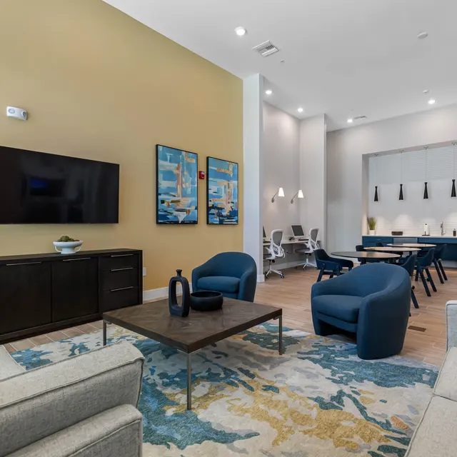 Modern Lounge Area A modern lounge area featuring comfortable seating, a large television mounted on the wall, and decorative artwork. The space is well-lit and includes a dining area with a table and chairs in the background.