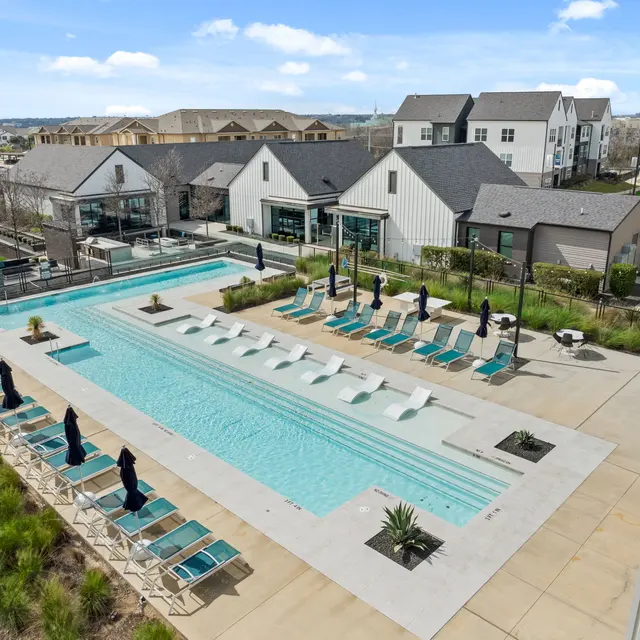 Aerial view of a modern apartment complex pool area, featuring a long swimming pool with sun loungers and umbrellas. Surrounding landscaping includes green plants and pathways.