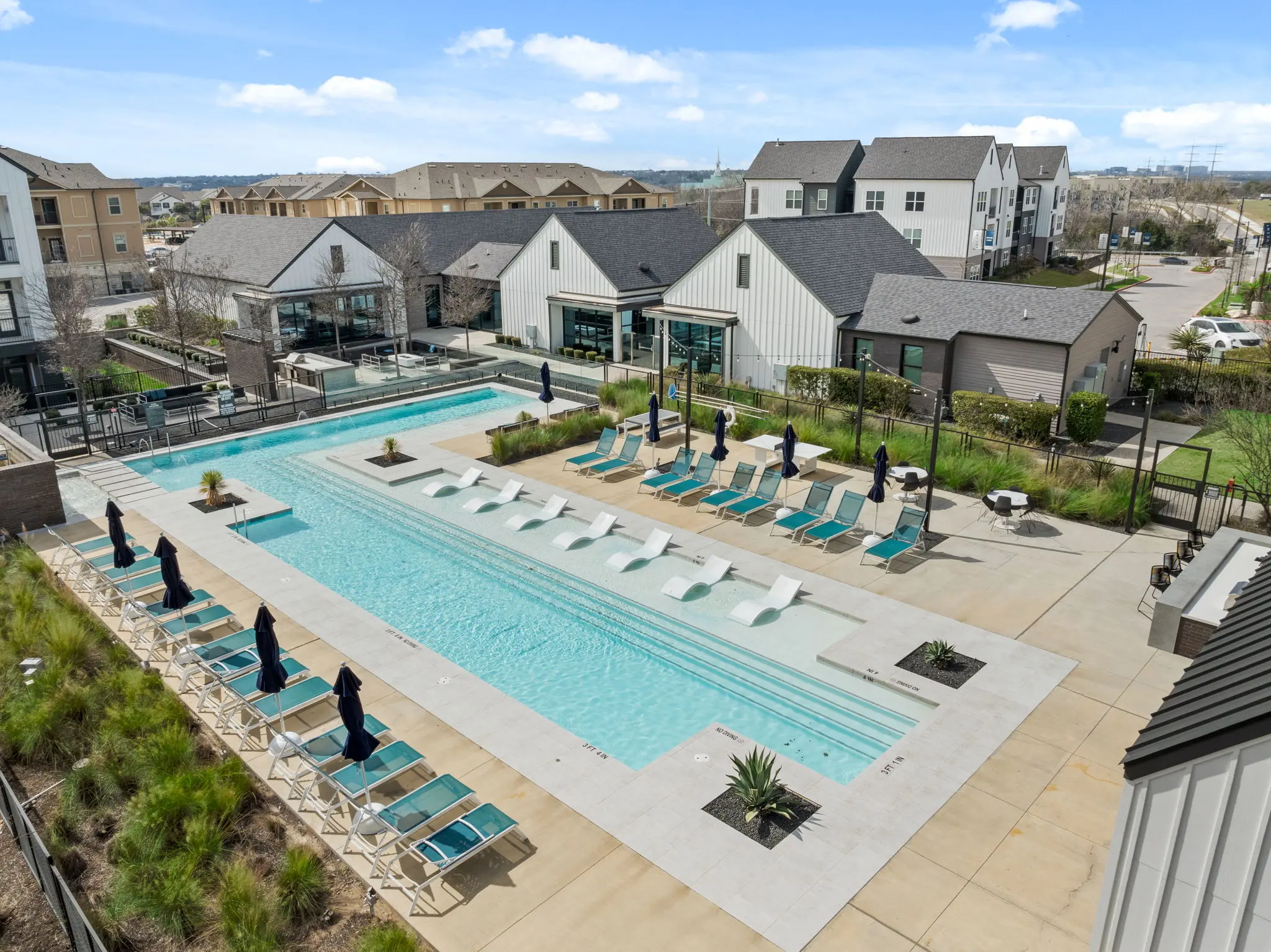 Aerial view of a modern apartment complex pool area, featuring a long swimming pool with sun loungers and umbrellas. Surrounding landscaping includes green plants and pathways.
