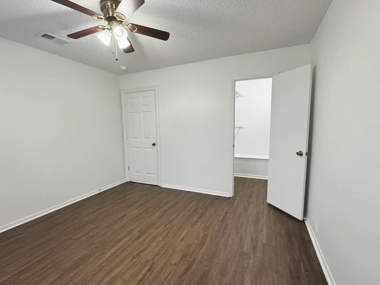 A spacious, empty room featuring a ceiling fan, light brown wooden flooring, and white walls. A closed door is visible to the left, while an open door leads to a small closet space on the right.