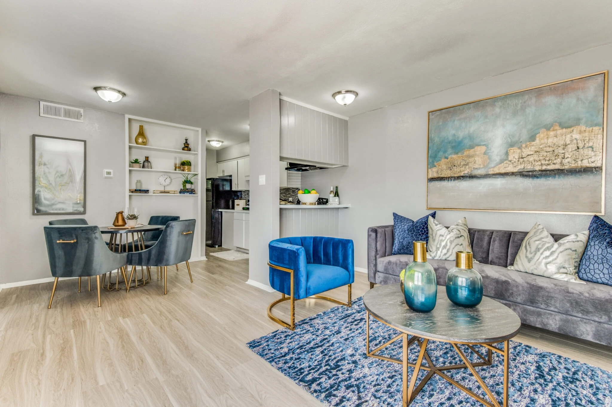 A modern living room with a dining area, featuring a gray couch, blue chairs, a round table, and decorative vases on a rug, showcasing a stylish and contemporary interior design.