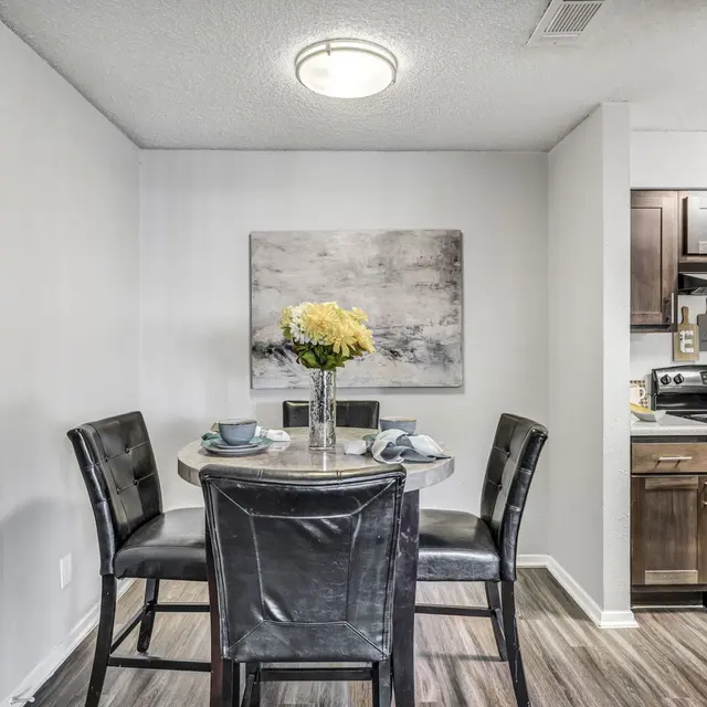 Modern dining area featuring a round table set with plates and flowers, surrounded by black chairs and an adjoining kitchen.