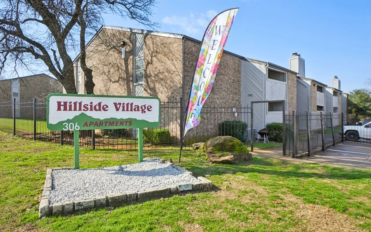 Sign for Hillside Village Apartments with colorful welcome banner and surrounding greenery.