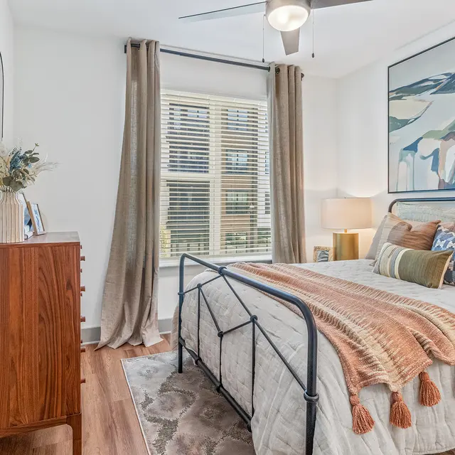A cozy and modern bedroom featuring a large bed with decorative pillows and a blanket, a wooden dresser, a window with light drapes, and a piece of abstract wall art.