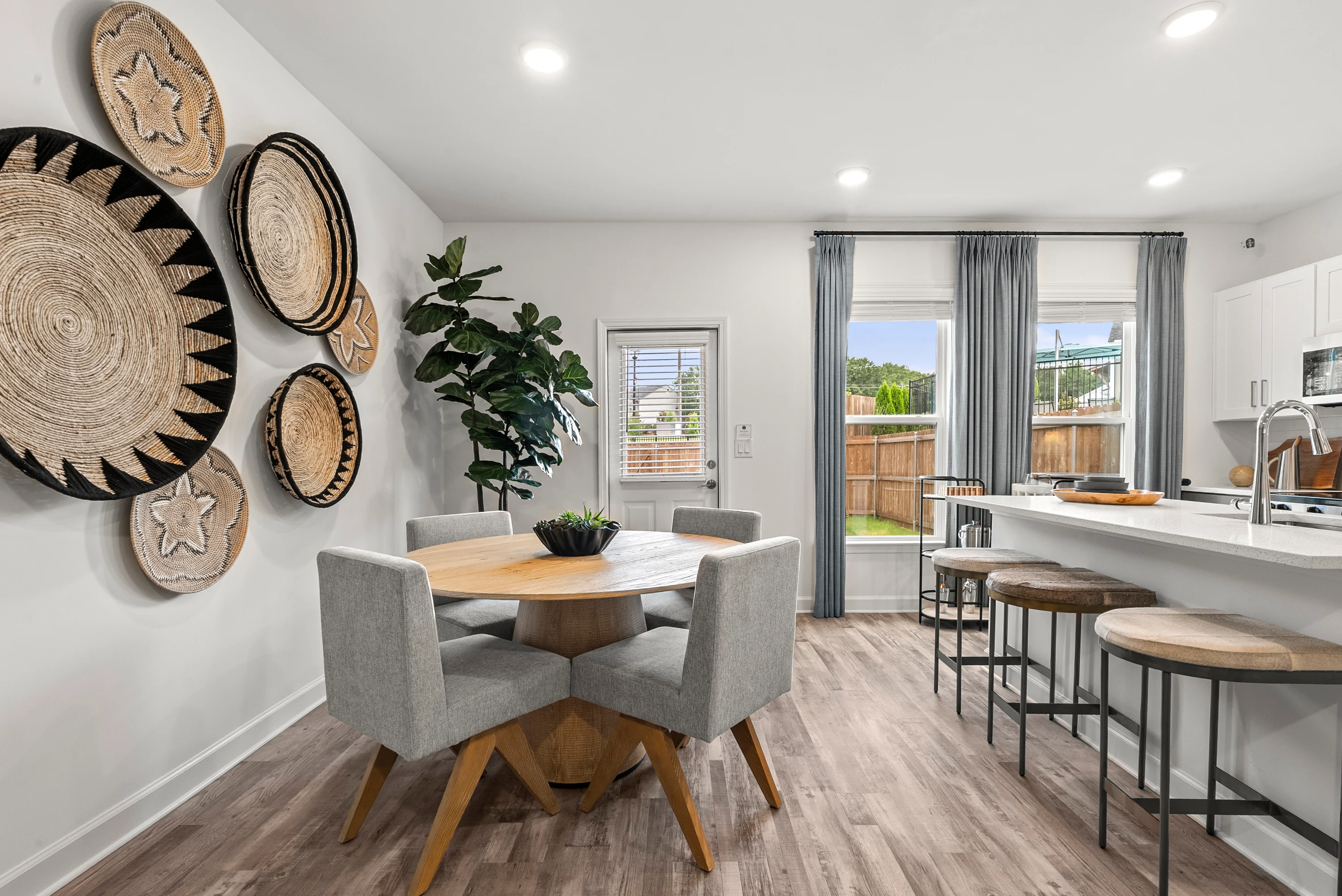 Modern Dining Space with Natural Light A modern dining area featuring a round wooden table with gray upholstered chairs. Decorative woven wall art adds style to the space. A large window offers natural light and a view of the patio.