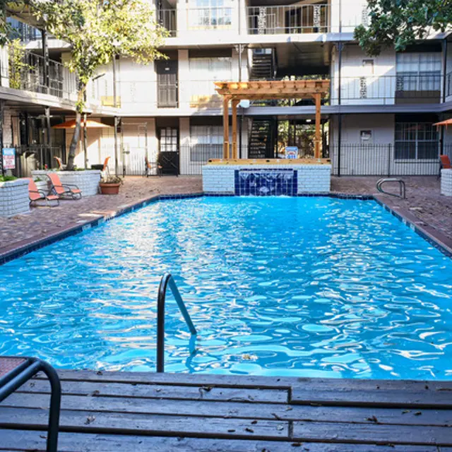 Watermill Apartments Pool Area A swimming pool surrounded by a patio area at a residential complex. There are lounge chairs positioned around the pool, and a wooden pergola is visible in the background. The pool is clear, reflecting the sunlight.
