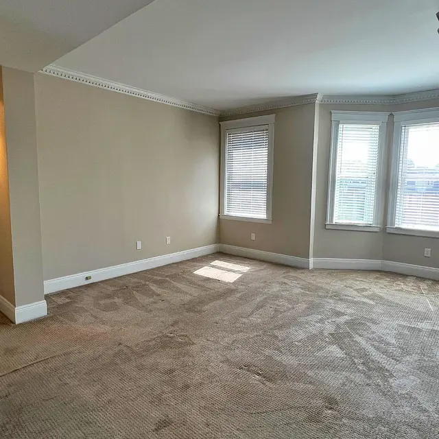An empty room with beige walls and a carpeted floor. Two windows with white blinds are visible, along with a closed door on the left. The room is well-lit with recessed lighting and has crown molding along the ceiling.