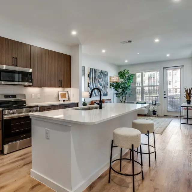 A modern kitchen with a large island, wooden cabinets, stainless steel appliances, and an open floor plan leading to a balcony.