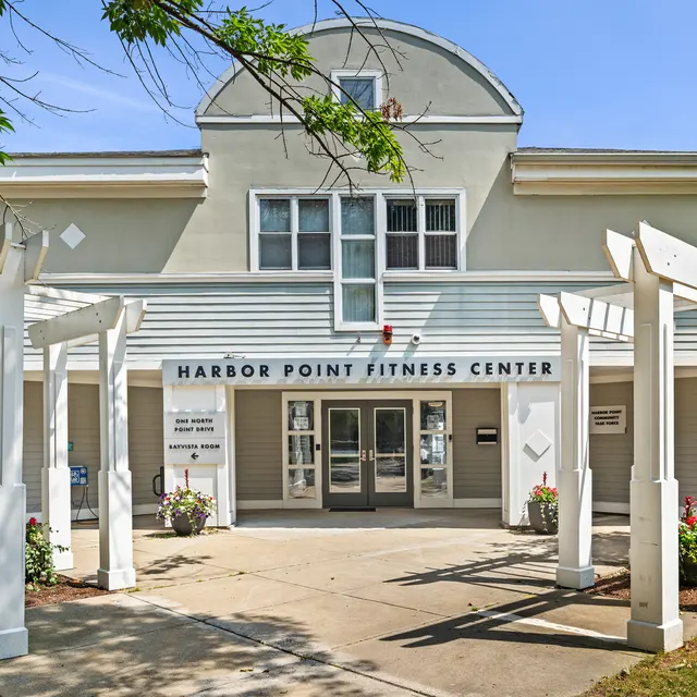Exterior view of the Harbor Point Fitness Center, featuring a light-colored building with large glass doors, surrounded by white wooden structures and green landscaping.