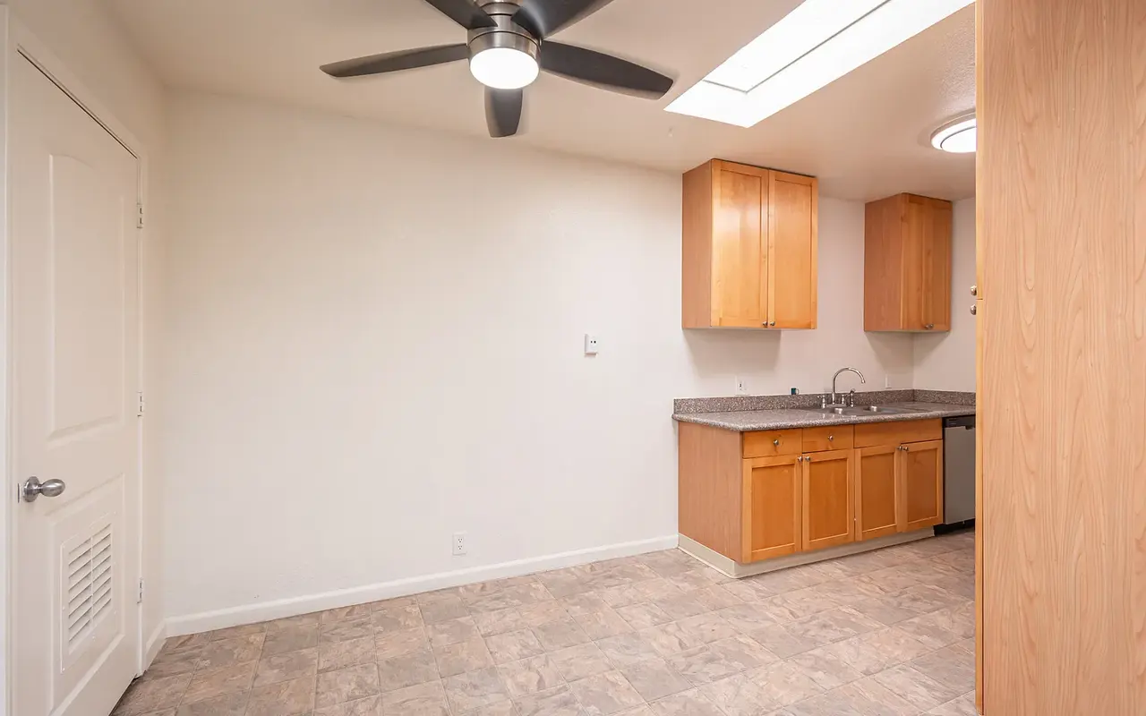 A minimally furnished kitchen with wooden cabinets, a countertop, and an open floor plan. The area has a ceiling fan and a skylight for natural lighting.