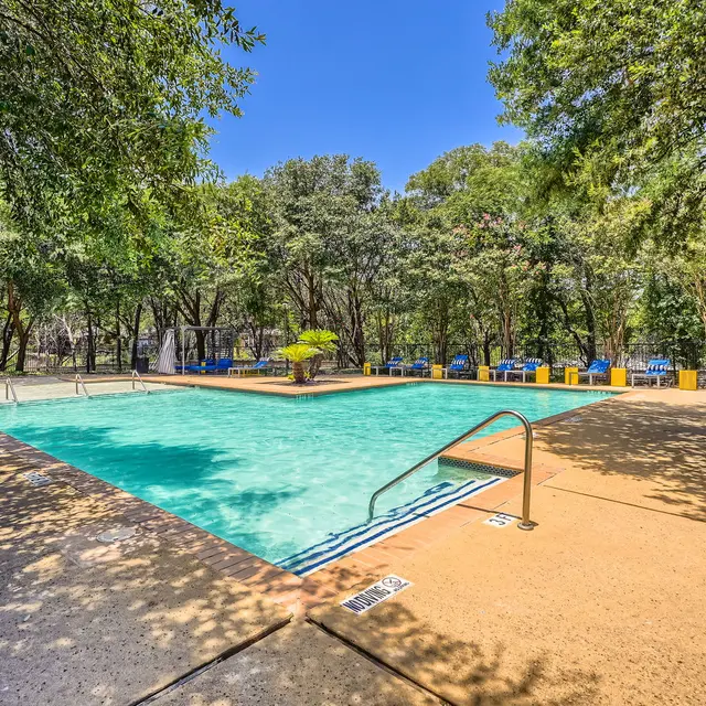 A bright and inviting swimming pool surrounded by greenery and trees, with lounge chairs nearby.