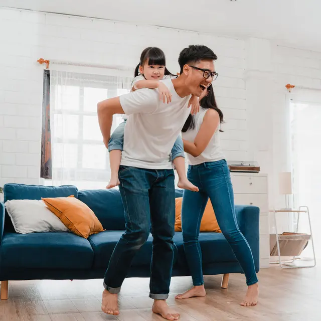 Family Fun in the Living Room A family having fun in a living room. A parent is giving a piggyback ride to a young girl, while another parent is smiling and posing next to them. There are soft furnishings and bright, natural light coming in through the windows.