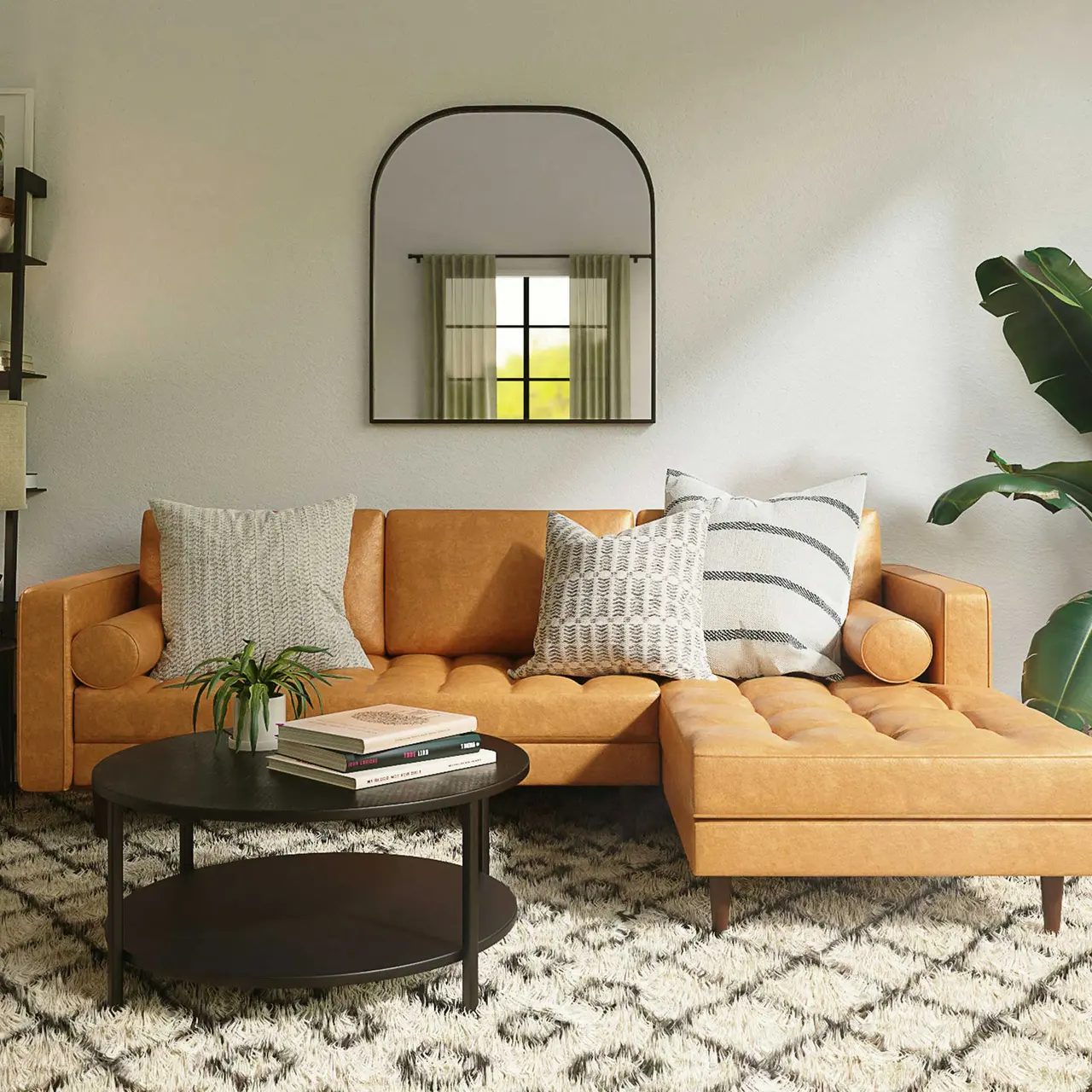 A modern living room featuring a stylish tan sectional sofa with various patterned cushions, a round black coffee table, a textured area rug, and a tall plant. A mirror hangs above the sofa, reflecting natural light from a window.