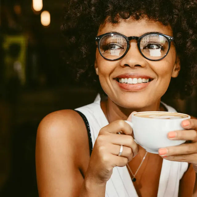 Woman Sipping Coffee A smiling woman with curly hair holding a cup of coffee in a cozy cafe setting.